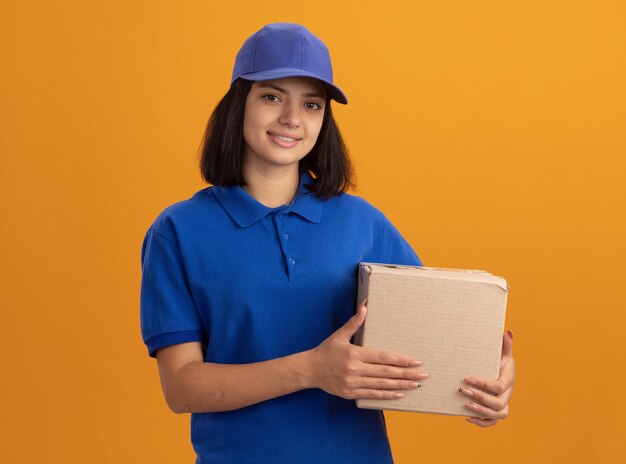 Junges Liefermädchen in der blauen Uniform und in der Kappe, die Pappkarton lächelnd freundlich stehend über orange Wand hält