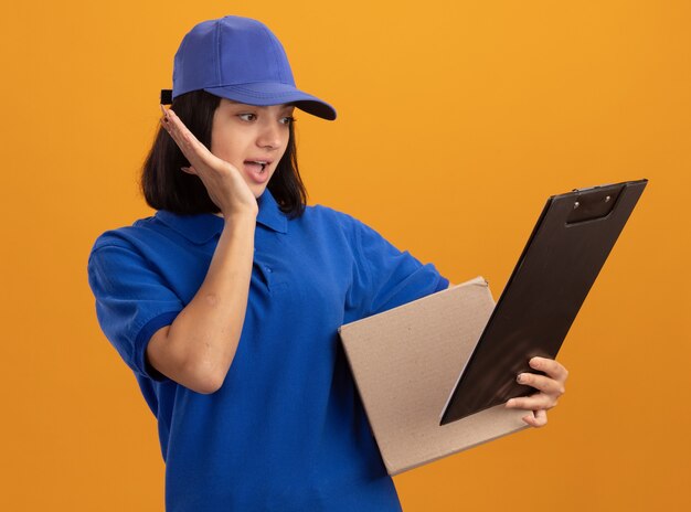 Junges Liefermädchen in der blauen Uniform und in der Kappe, die Pappkarton und Zwischenablage hält, die es überrascht betrachten, über orange Wand zu stehen