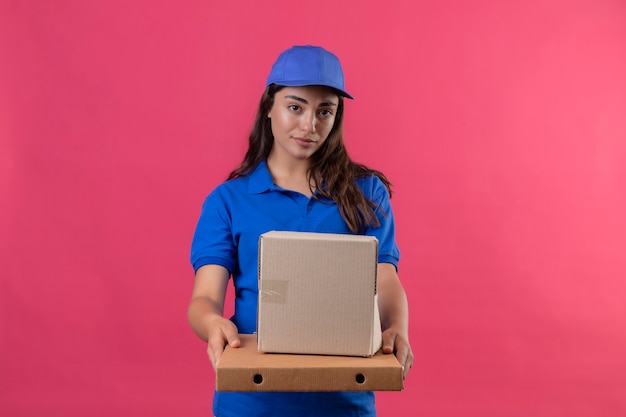 Junges Liefermädchen in der blauen Uniform und in der Kappe, die Pappkartons hält, die Kamera mit ernstem sicherem Ausdruck auf Gesicht stehen, das über rosa Hintergrund steht