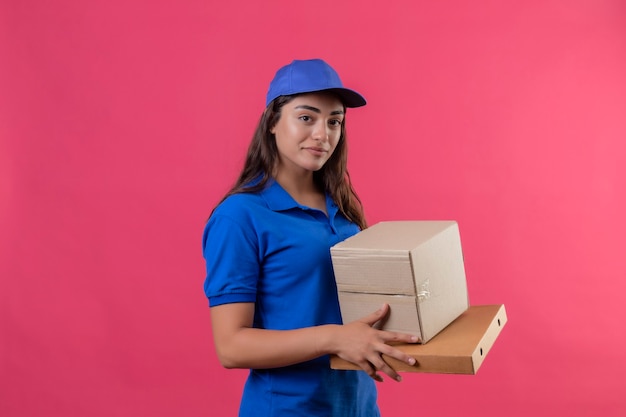 Junges Liefermädchen in der blauen Uniform und in der Kappe, die Pappkartons hält, die zuversichtlich positiv und glücklich über rosa Hintergrund lächeln