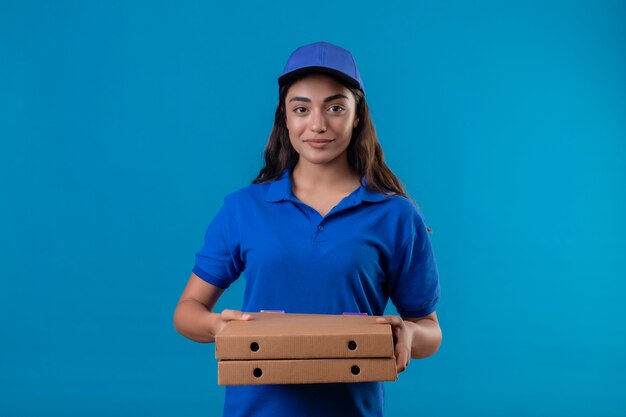 Junges liefermädchen in der blauen uniform und in der kappe, die pizzakästen betrachten, die kamera betrachten, die zuversichtlich glücklich und positiv steht über blauem hintergrund