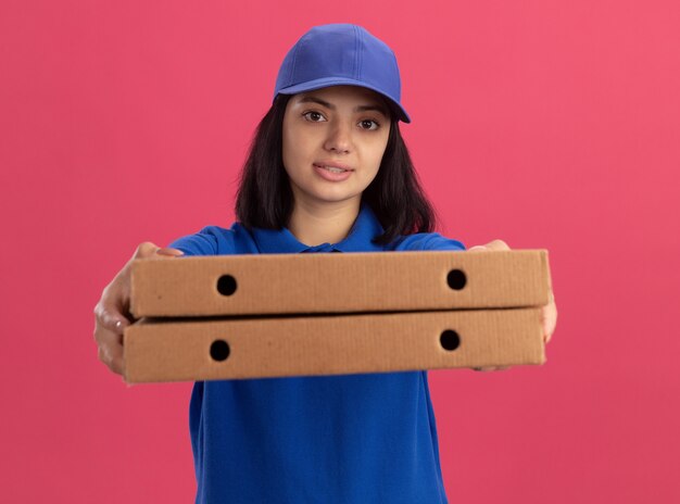 Junges Liefermädchen in der blauen Uniform und in der Kappe, die Pizzaschachteln hält, die sicher aussehend stehen über rosa Wand strecken