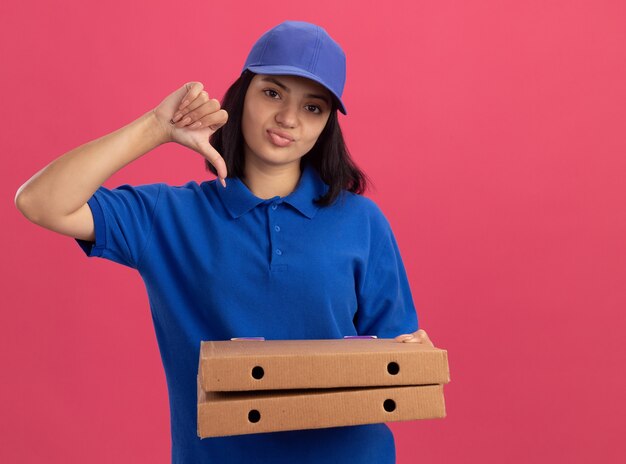 Junges Liefermädchen in der blauen Uniform und in der Kappe, die Pizzaschachteln halten, missfiel, Daumen nach unten stehend über rosa Wand stehend