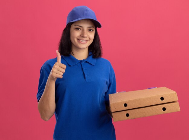 Junges Liefermädchen in der blauen Uniform und in der Kappe, die Pizzaschachteln lächelnd mit glücklichem Gesicht zeigt, das Daumen oben steht über rosa Wand