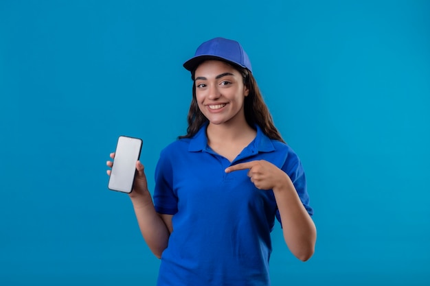 Junges Liefermädchen in der blauen Uniform und in der Kappe, die Smartphone zeigt, zeigt mit dem Finger, der freundlich über blauem Hintergrund stehend lächelt