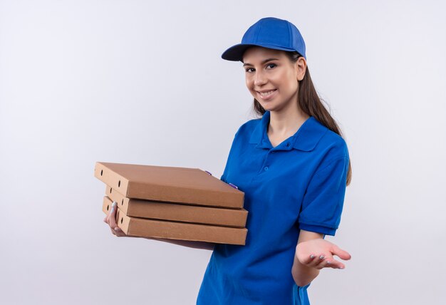 Junges Liefermädchen in der blauen Uniform und in der Kappe, die Stapel von Pizzaschachteln hält, die Kamera betrachten, die fröhlich den Arm ausstreckt und um Zahlung bittet