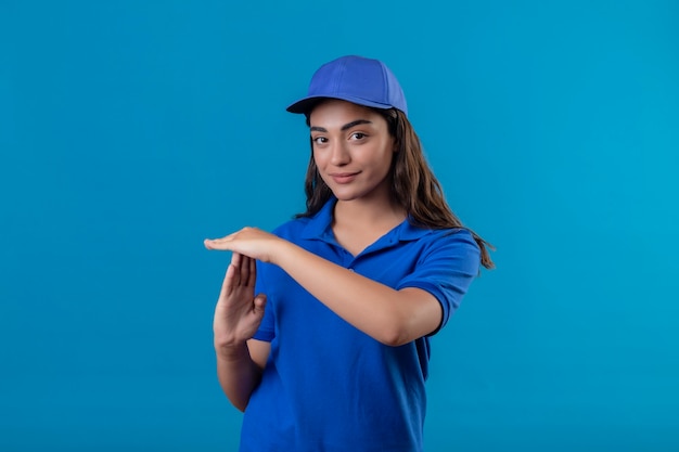 Junges Liefermädchen in der blauen Uniform und in der Kappe, die zuversichtlich schauen, Auszeitgeste mit Händen zu machen, die über blauem Hintergrund stehen