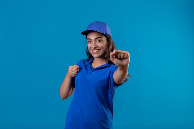 Junges Liefermädchen in der blauen Uniform und in der Kappe lächelnd, die fröhlich posiert wie ein Boxer mit Fäusten, die über blauem Hintergrund stehen