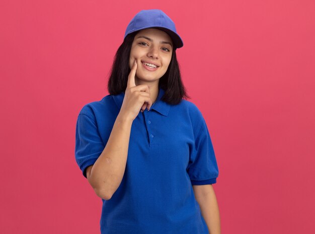 Junges Liefermädchen in der blauen Uniform und in der Kappe lächelnd mit glücklichem Gesicht mit Finger an ihrer Wange, die über rosa Wand steht