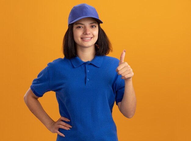 Junges Liefermädchen in der blauen Uniform und in der lächelnden Kappe, die Tumben oben stehen über orange Wand zeigt