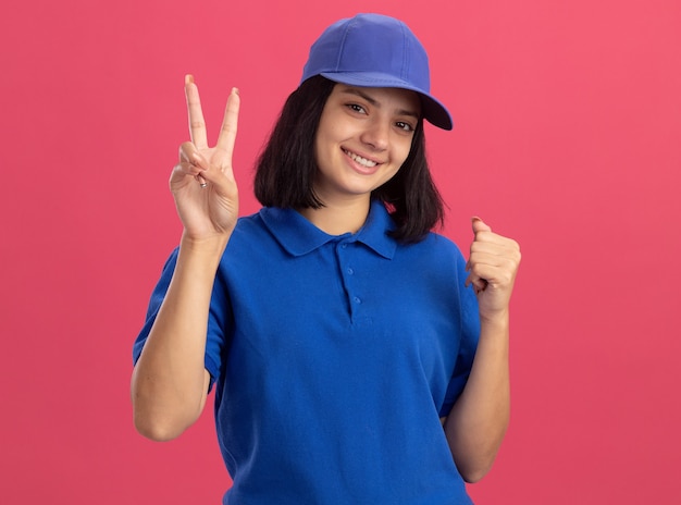 Junges Liefermädchen in der blauen Uniform und in der lächelnden Kappe, die V-Zeichen zeigt, die Faust glücklich und positiv ballen, die über rosa Wand stehen