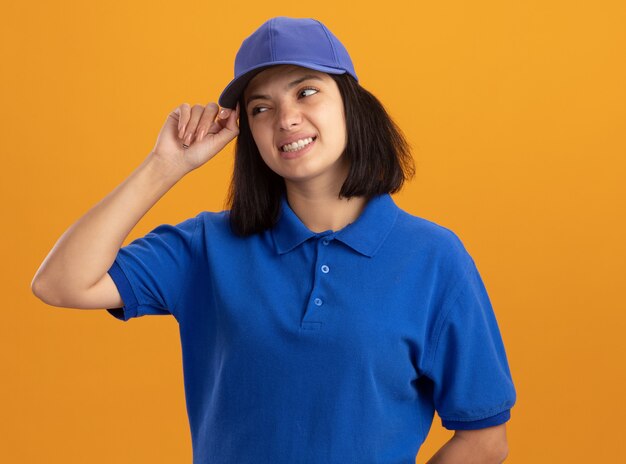 Junges Liefermädchen in der blauen Uniform und in der Mütze, die lächelnd verwirrt über orange Wand stehend schaut