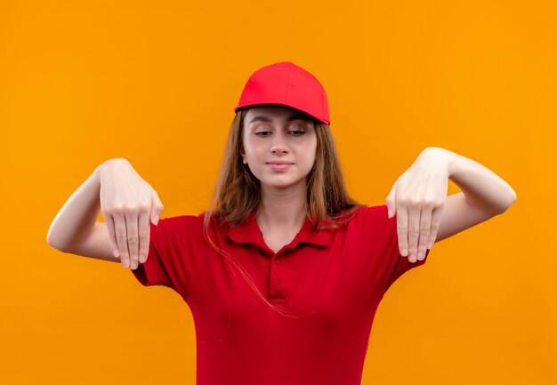 Junges Liefermädchen in der roten Uniform, die mit Händen unten zeigt und auf isolierte orange Wand unten schaut