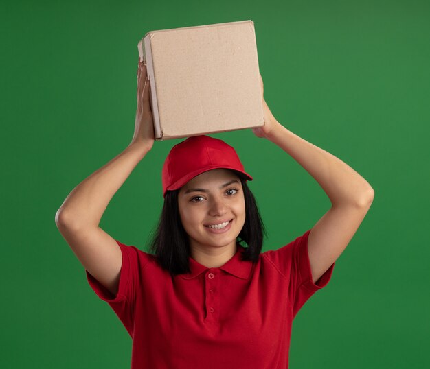 Junges Liefermädchen in der roten Uniform und in der Kappe, die Pappkarton über ihrem Kopf hält lächelnd freundlich stehend über grüner Wand