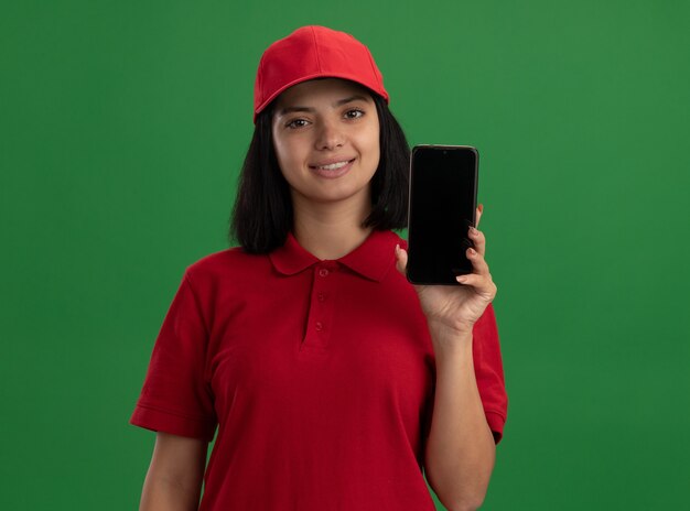 Junges Liefermädchen in der roten Uniform und in der Kappe, die Smartphone mit Lächeln auf Gesicht über grüner Wand stehend zeigt