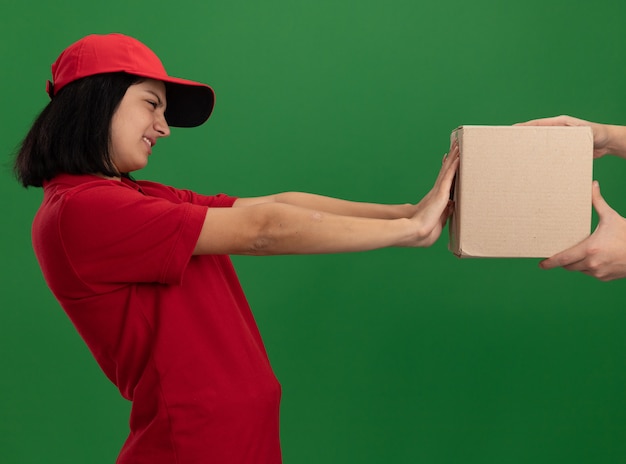 Kostenloses Foto junges liefermädchen in roter uniform und kappe, die sich weigern, ein papierpaket zu nehmen, das über grüner wand steht