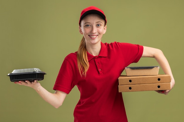 Junges Liefermädchen in roter Uniform und Mütze, das Pizzakartons und Lebensmittelpakete hält