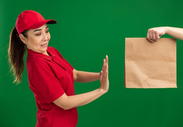 Junges Liefermädchen in roter Uniform und Mütze, das sich weigert, ein Papierpaket zu erhalten, das über einer grünen Wand steht?