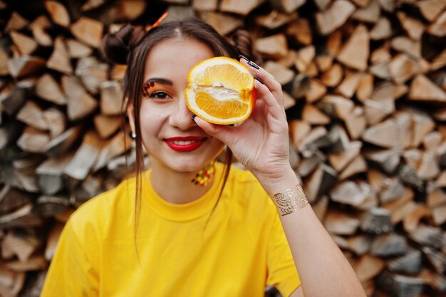 Junges lustiges Mädchen mit heller Make-up-Abnutzung auf gelbem Hemd hält ein Stück Orange vor Holzhintergrund