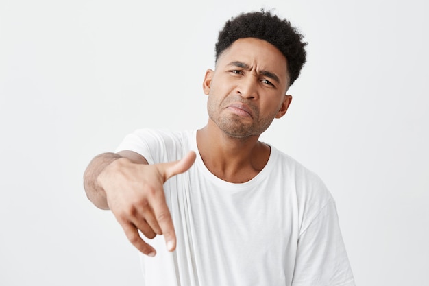 Junges lustiges schönes schwarzhäutiges Männchen mit Afro-Frisur im lässigen weißen T-Shirt, das in der Kamera mit Hand zeigt, lustiges Gesicht macht, das für Foto mit Freunden auf Partei aufwirft.