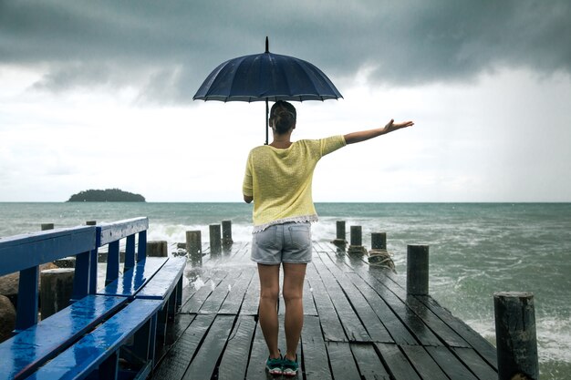 junges Mädchen am Pier mit Regenschirm steht mit dem Rücken zum Meer