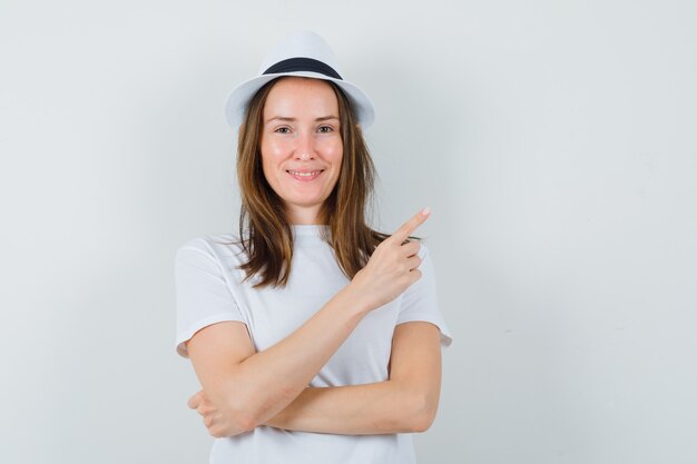 Junges Mädchen, das an der oberen rechten Ecke in weißem T-Shirt, Hut zeigt und zuversichtlich schaut, Vorderansicht.