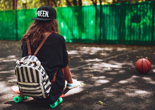 Junges Mädchen, das auf Kunststoff orange Penny Shortboard auf Asphalt in Kappe sitzt