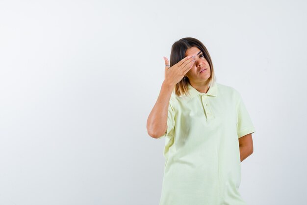 Junges Mädchen, das Auge mit Hand bedeckt, Hand hinter Taille im T-Shirt hält und zuversichtlich schaut. Vorderansicht.