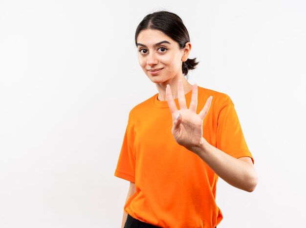 Junges Mädchen, das das lächelnde orange T-Shirt zeigt und mit den Fingern oben Nummer vier über der weißen Wand stehend zeigt