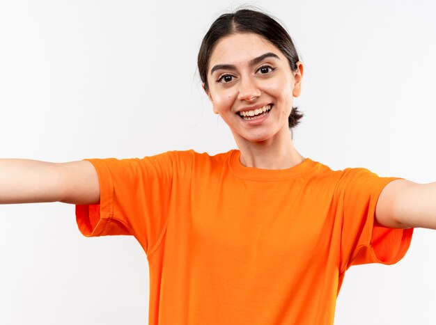 Junges Mädchen, das das orange T-Shirt trägt, das freundlich lächelt und Begrüßungsgeste mit den Händen macht, die über weißer Wand stehen