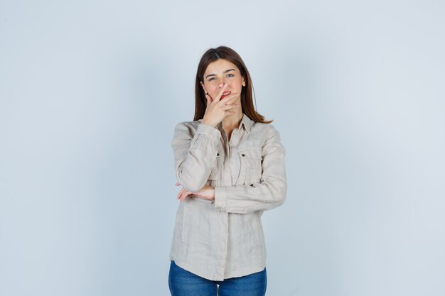 Junges Mädchen, das den Mund bedeckt, während es in beigefarbenem Hemd, Jeans lächelt und fröhlich aussieht. Vorderansicht.