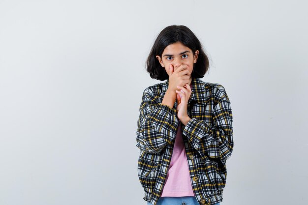 Junges Mädchen, das den Mund mit den Händen im roten T-Shirt und in der Jeansjacke bedeckt und überrascht schaut. Vorderansicht.