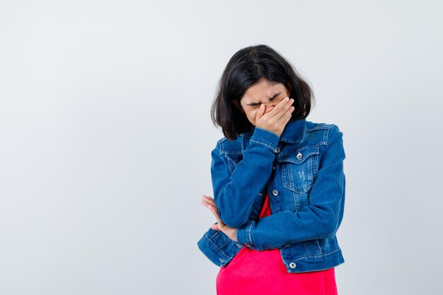 Junges Mädchen, das den Mund mit der Hand bedeckt, in rotem T-Shirt und Jeansjacke gähnt und müde aussieht