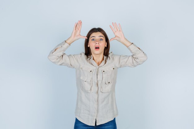 Junges Mädchen, das die Hände in der Nähe des Kopfes als Ohren hebt, den Mund in beigefarbenem Hemd, Jeans weit offen hält und überrascht aussieht, Vorderansicht.