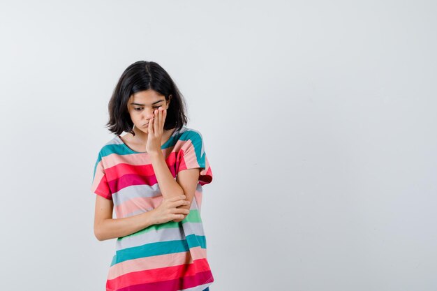 Junges Mädchen, das die Hand auf die Wange legt, während es in denkender Pose in bunt gestreiftem T-Shirt steht und nachdenklich aussieht. Vorderansicht.