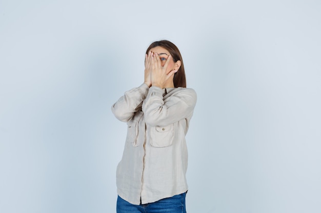 Junges Mädchen, das durch die Finger in beigefarbenem Hemd, Jeans schaut und sich schämt, Vorderansicht.