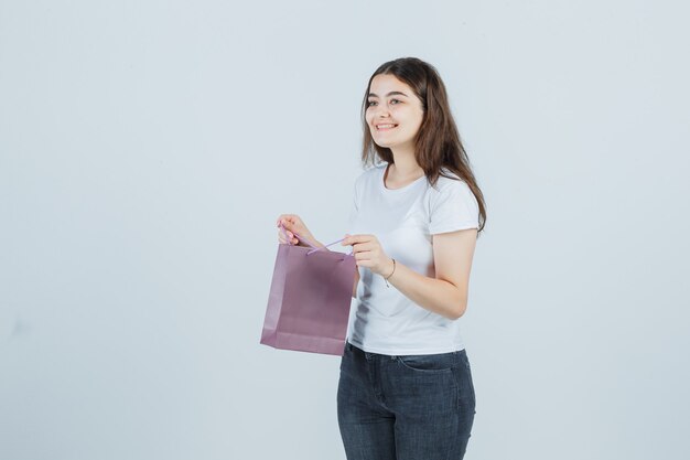 Junges Mädchen, das eine Papiertüte im T-Shirt, in den Jeans hält und fröhlich, Vorderansicht schaut.