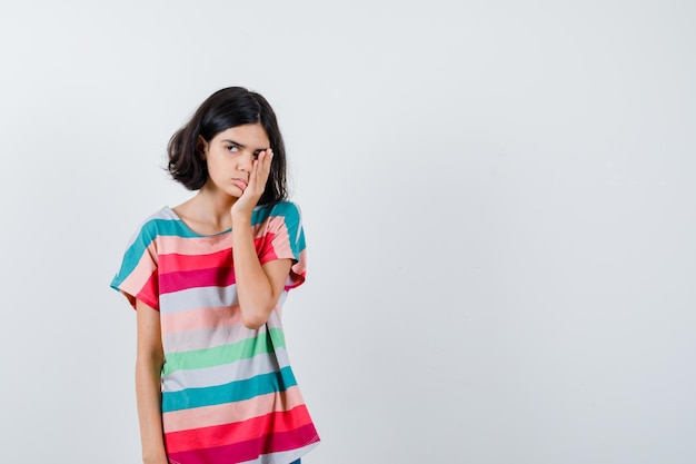 Junges Mädchen, das einen Teil des Gesichts mit der Hand in einem bunt gestreiften T-Shirt bedeckt und erschöpft aussieht. Vorderansicht.