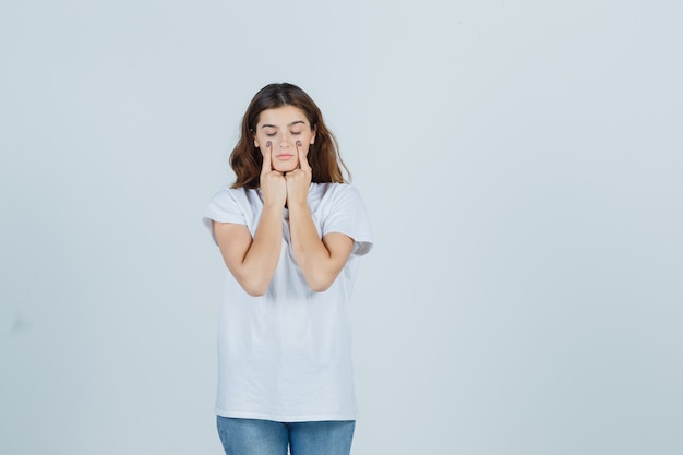 Junges Mädchen, das Finger auf Augenlidern im weißen T-Shirt hält und vernünftig aussieht. Vorderansicht.