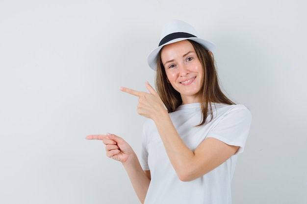 Junges Mädchen, das Finger auf die linke Seite im weißen T-Shirt, im Hut zeigt und lustig schaut. Vorderansicht.