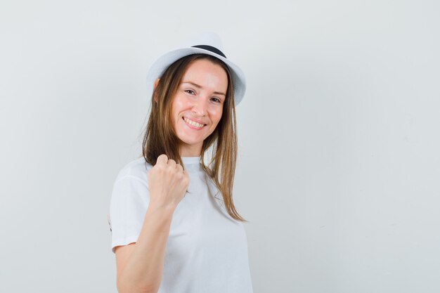 Junges Mädchen, das geballte Faust in weißem T-Shirt, Hut zeigt und Glück schaut. Vorderansicht.