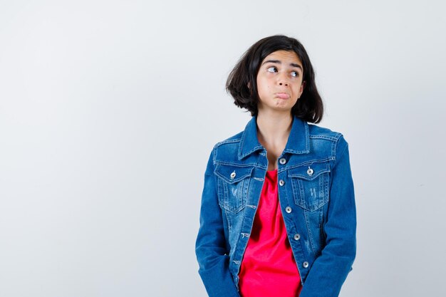 Junges Mädchen, das gerade steht und in die Kamera posiert, in rotem T-Shirt und Jeansjacke wegschaut und glücklich aussieht