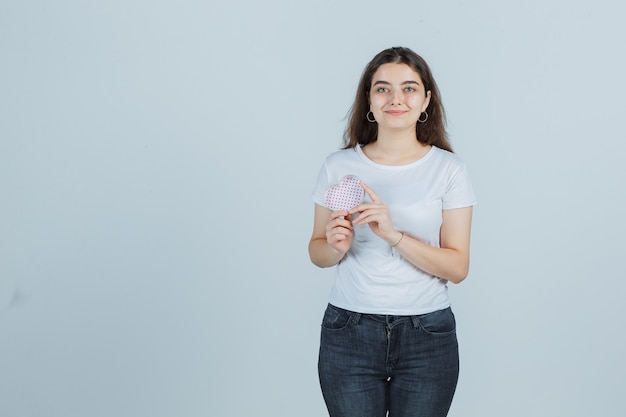 Junges Mädchen, das Geschenkbox hält, während Box in T-Shirt, Jeans betrachtet und fröhlich, Vorderansicht schaut.