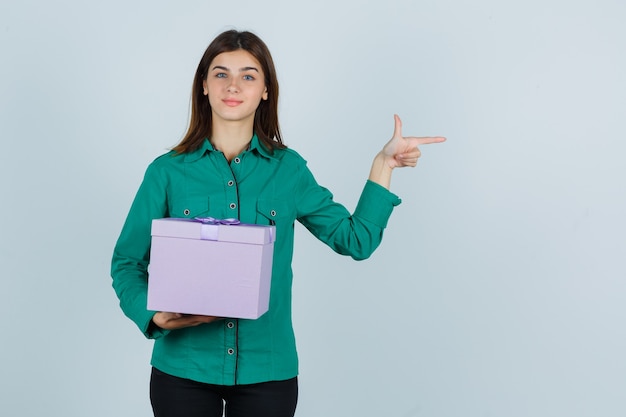 Junges Mädchen, das Geschenkbox hält, zeigt rechts mit Zeigefinger in grüner Bluse, schwarze Hose und schaut fröhlich, Vorderansicht.