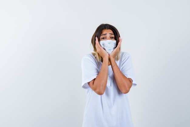 Junges Mädchen, das Gesicht auf Händen im weißen T-Shirt und in der Maske kissen und besorgt, Vorderansicht schaut.