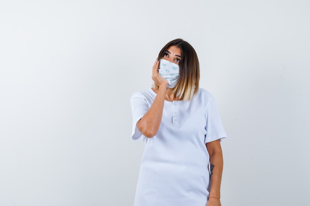 Junges Mädchen, das Gesicht auf Hand im weißen T-Shirt, Maske und nachdenklich aussehend, Vorderansicht kissen.