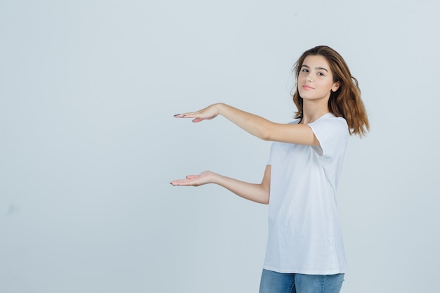 Kostenloses Foto junges mädchen, das größenzeichen in t-shirt, jeans zeigt und zuversichtlich schaut. vorderansicht.