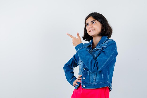 Junges Mädchen, das Hand an der Taille hält, wegschaut, in rotem T-Shirt und Jeansjacke nach links zeigt und nachdenklich aussieht. Vorderansicht.