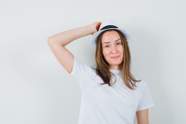 Junges Mädchen, das Hand auf Kopf im weißen T-Shirt, Hut hält und zuversichtlich schaut. Vorderansicht.