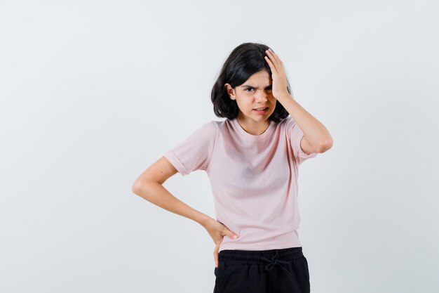 Junges Mädchen, das Hand auf Kopf setzt, während Hand auf Taille in rosa T-Shirt und in schwarzen Hosen hält und wütend schaut